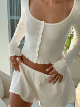 Close-up of a woman wearing a white knit button-front top and matching shorts by Florence by Mills. Millie Bobby Brown wears red knitted crop top and trousers