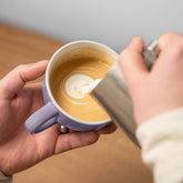 Close-up of hands pouring milk to create latte art in a purple mug. Millie Bobby Brown wears red knitted crop top and trousers