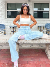 Young woman in a casual outfit with white crop top, light jeans, and pink shoes, sitting on an ornate bench. Millie Bobby Brown wears red knitted crop top and trousers